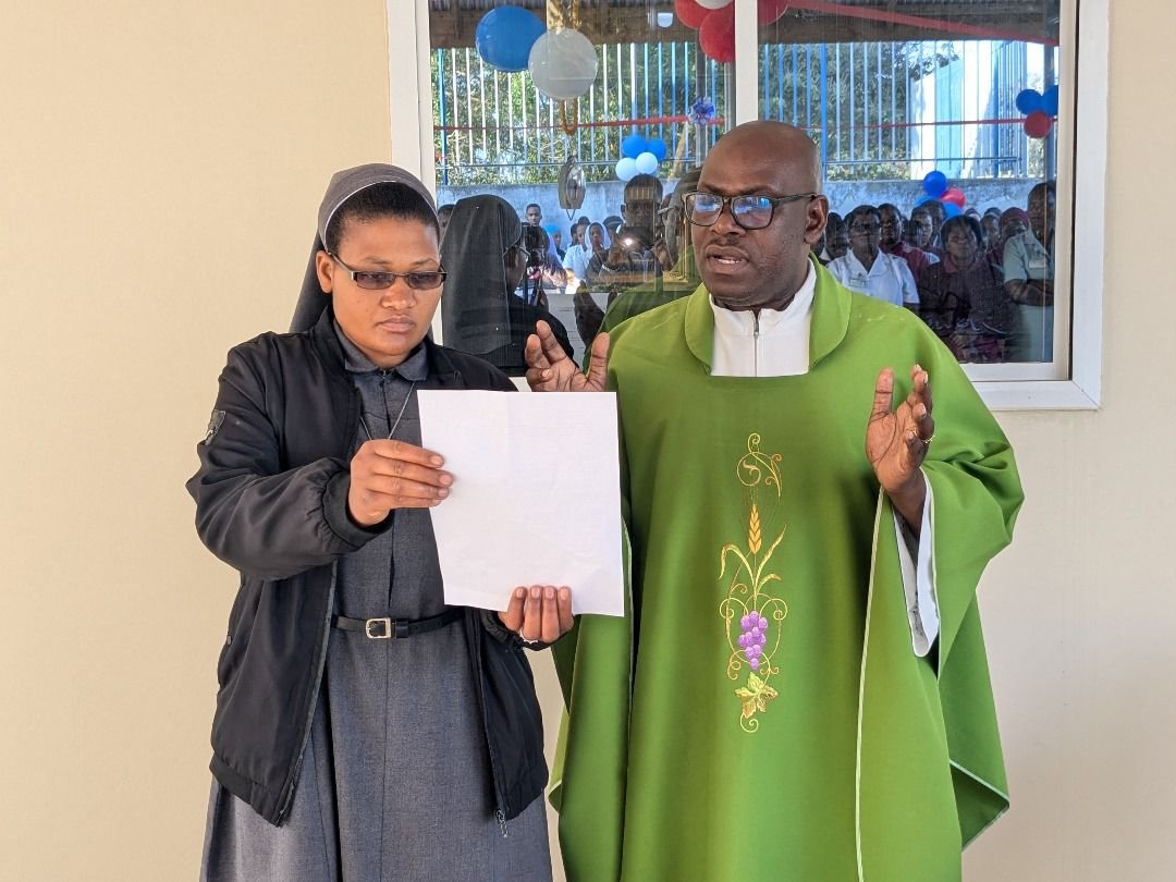 Una suora legge da un foglio accanto a un sacerdote con la casula verde, davanti a molte persone