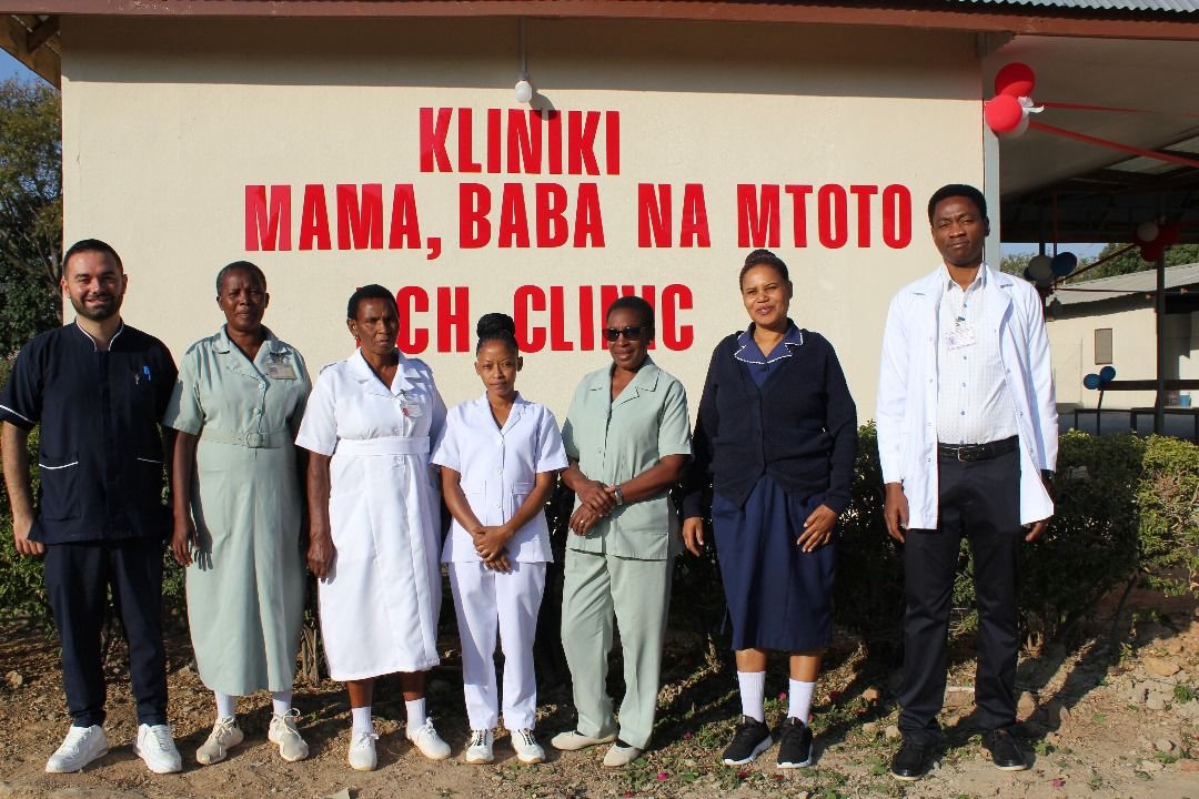 Sette persone del personale in divisa davanti al muro della clinica con la scritta rossa Kliniki Mama, Baba na Mtoto RCH Clinic