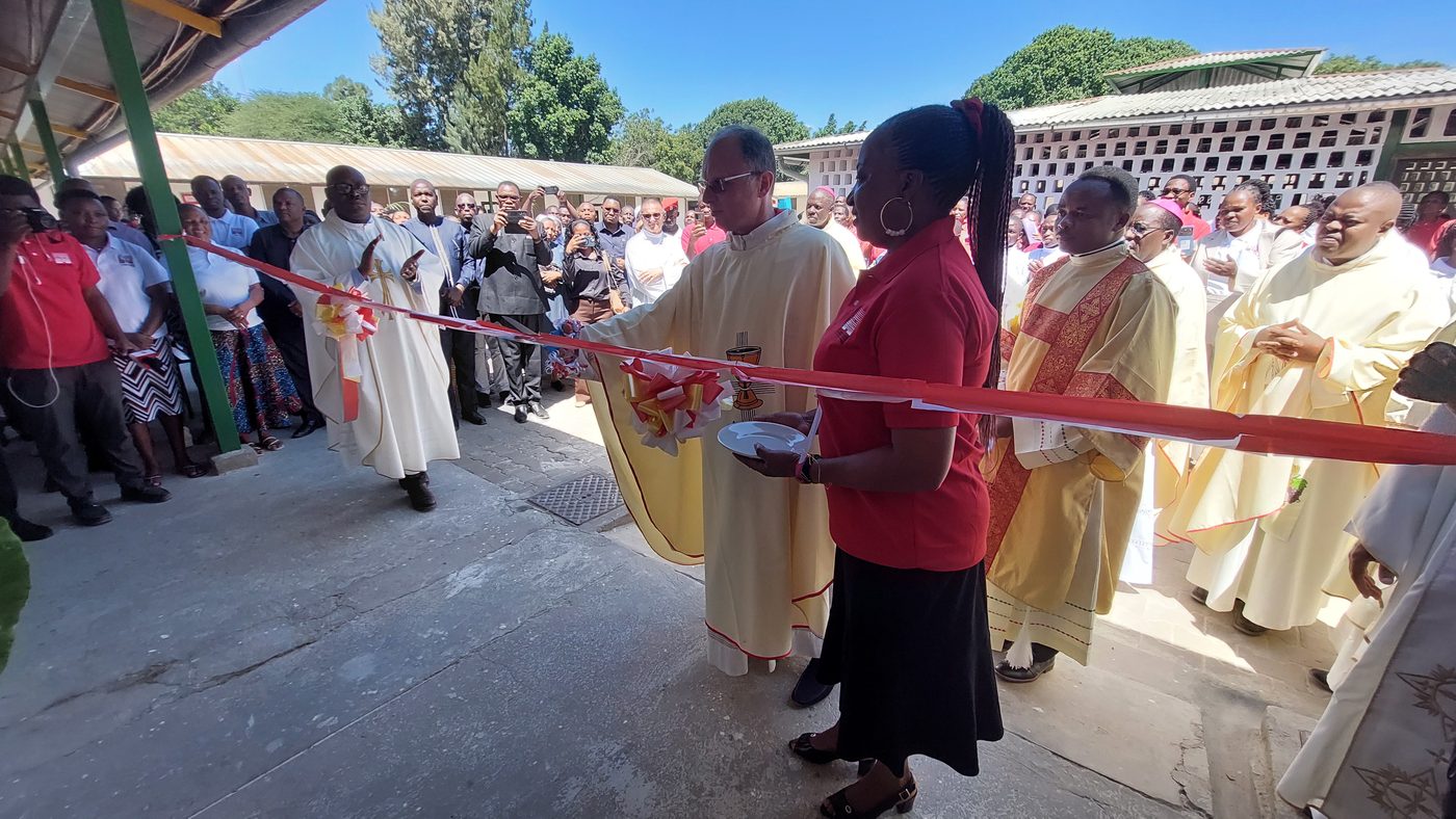 Un sacerdote con la casula e una donna con la maglia rossa davanti al nastro rosso, tra molte persone, all’ingresso del reparto